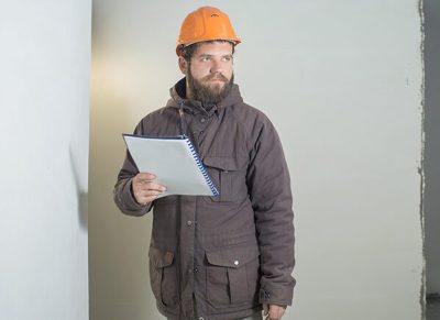 a young man with a beard is standing in an orange construction helmet with a folder of papers in his hands.